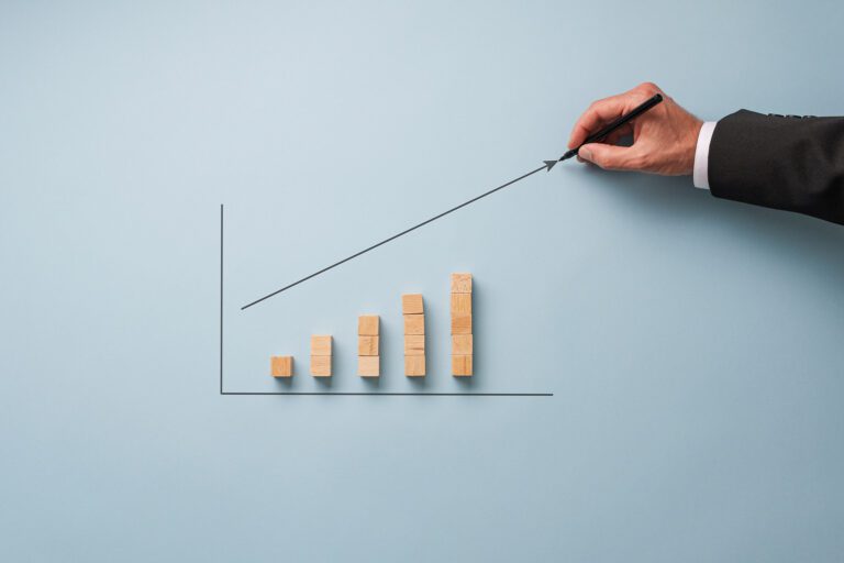 A hand in a suit holding a pen draws an upward-sloping arrow on a bar graph. The graph consists of five vertical wooden bars of increasing height from left to right, set against a light blue background. This image symbolizes growth and progress, typically associated with business or financial success.