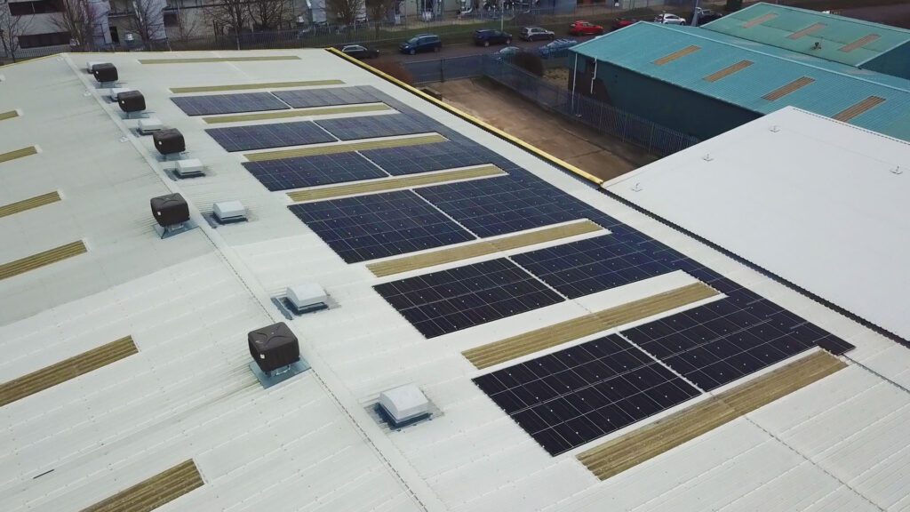 Rooftop of a large industrial building equipped with rows of solar panels covering significant space. Ventilation units are interspersed among the panels, showcasing functional rooftop use. The building is situated in a commercial area, surrounded by other structures and parked vehicles in the background, emphasizing urban renewable energy implementation.
