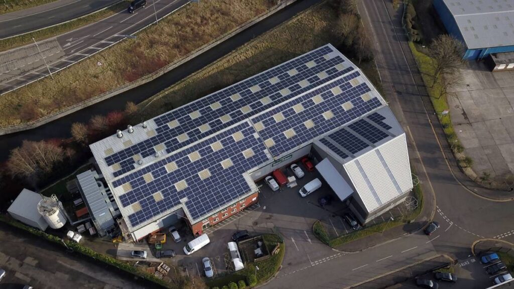 Aerial view of an industrial building with a roof covered in solar panels, parked vehicles, and a canal nearby.