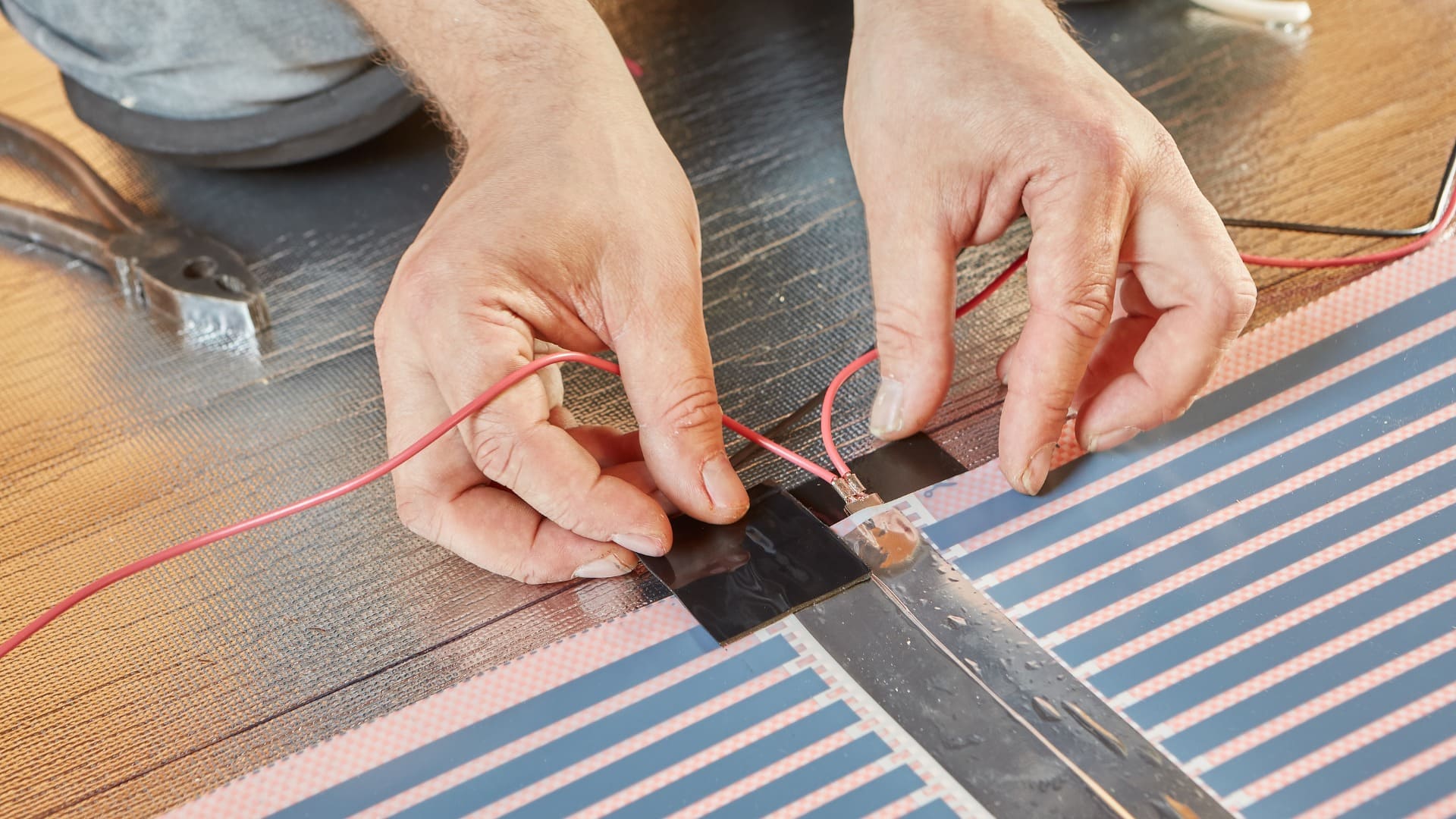 Close-up of hands working on electrical wiring, connecting red wire to part of an underfloor heating system. The wire is secured with black tape, with pliers visible in the background, showing the intricacies of home improvement