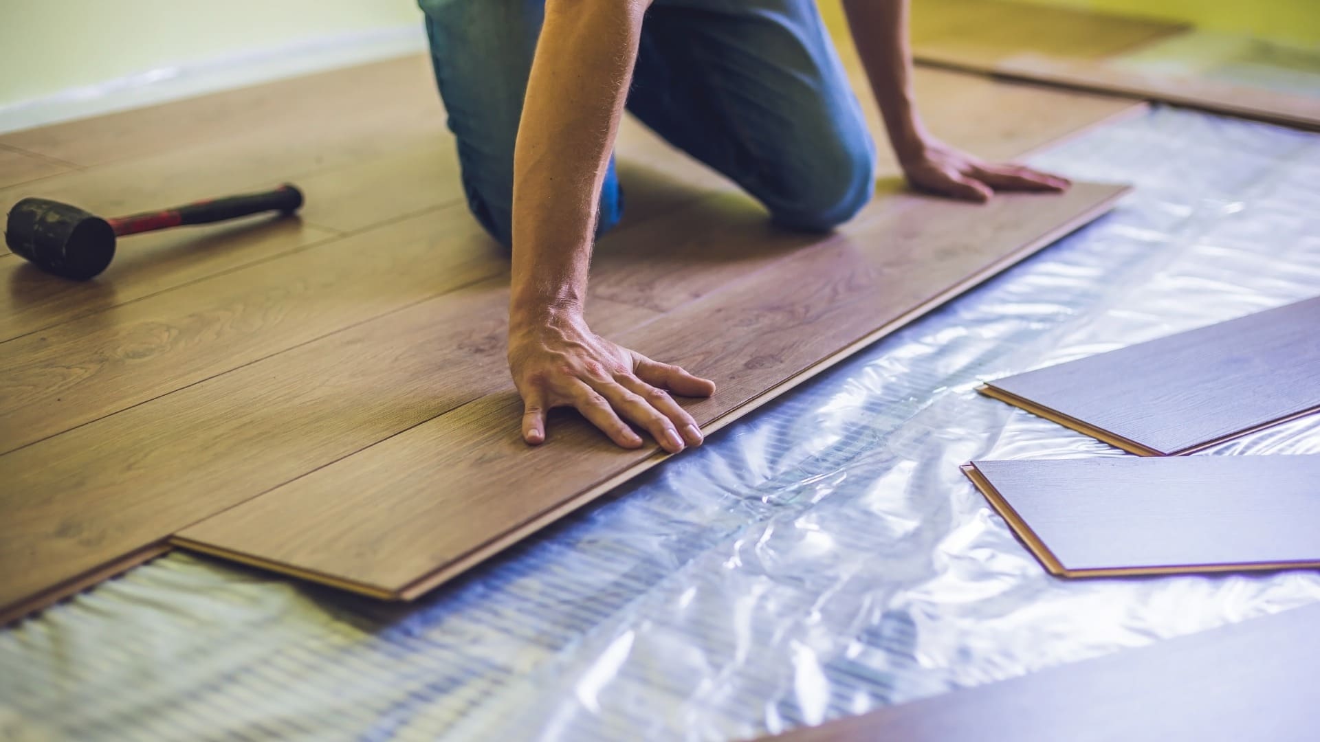 Person installing wooden flooring, kneeling to position wooden planks over a plastic moisture barrier, with a rubber mallet nearby and additional planks in the foreground.
