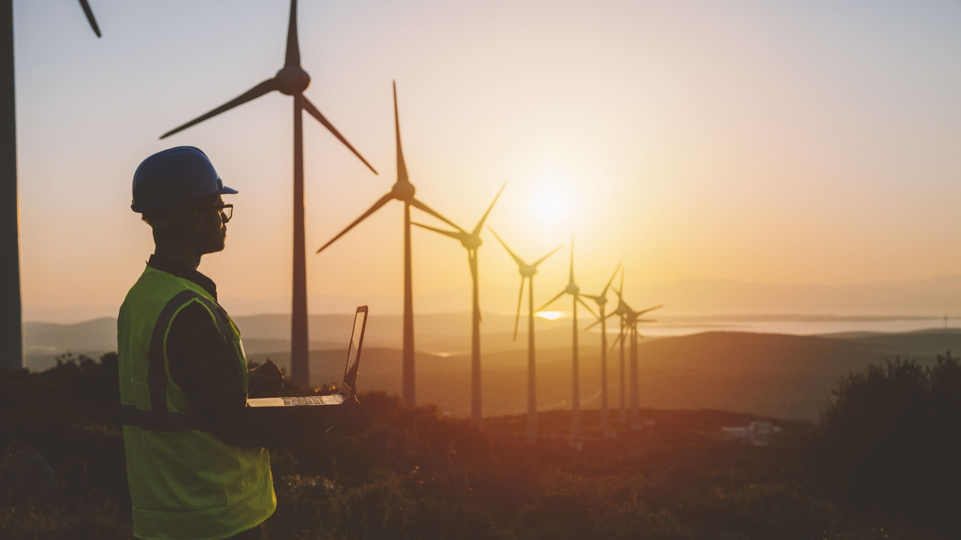 Solar panels in rows with two wind turbines spinning in the background, set against a warm sunrise or sunset.