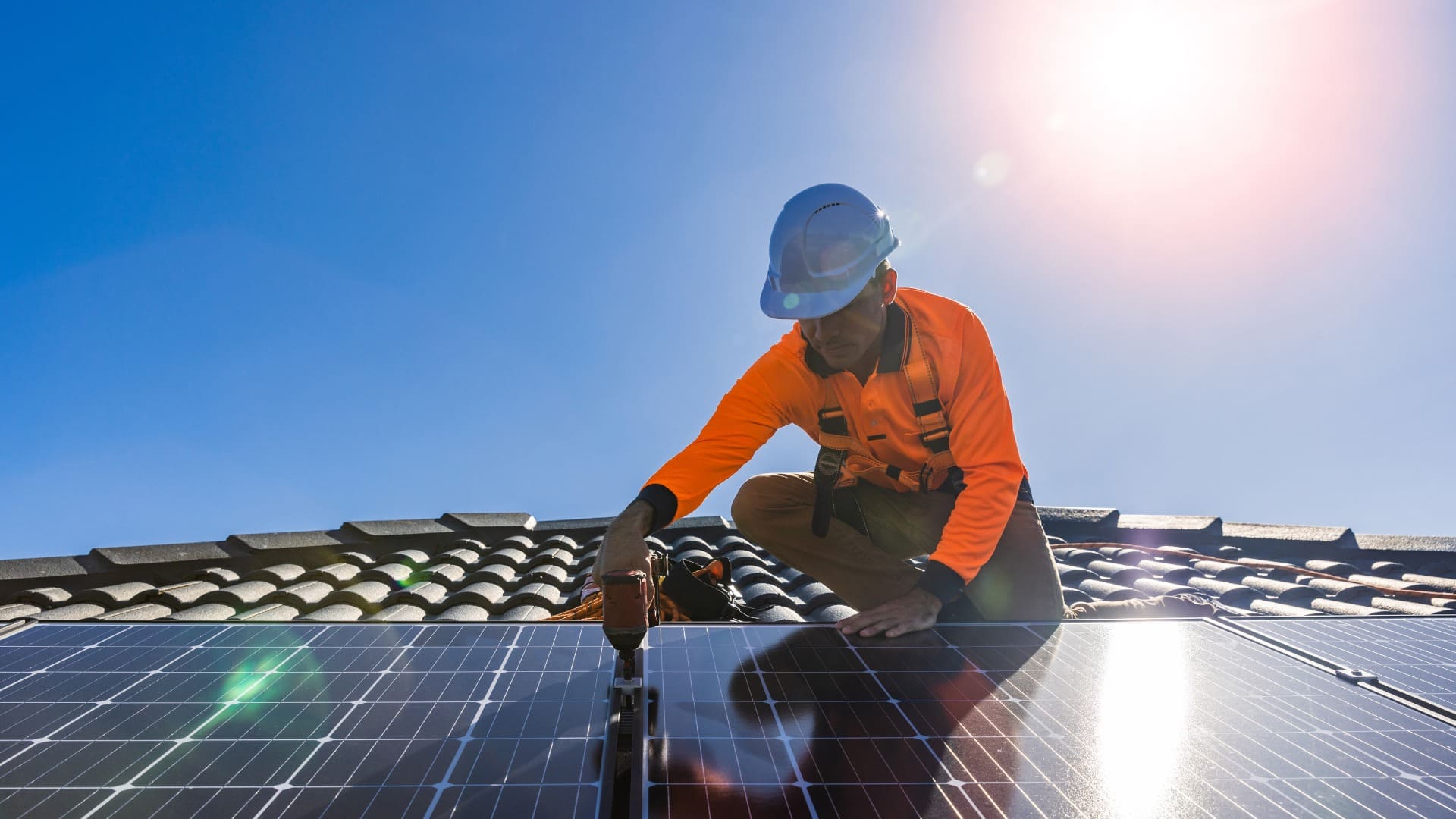 Worker in safety gear installing solar panels on tiled roof under bright blue sky, using power tools.