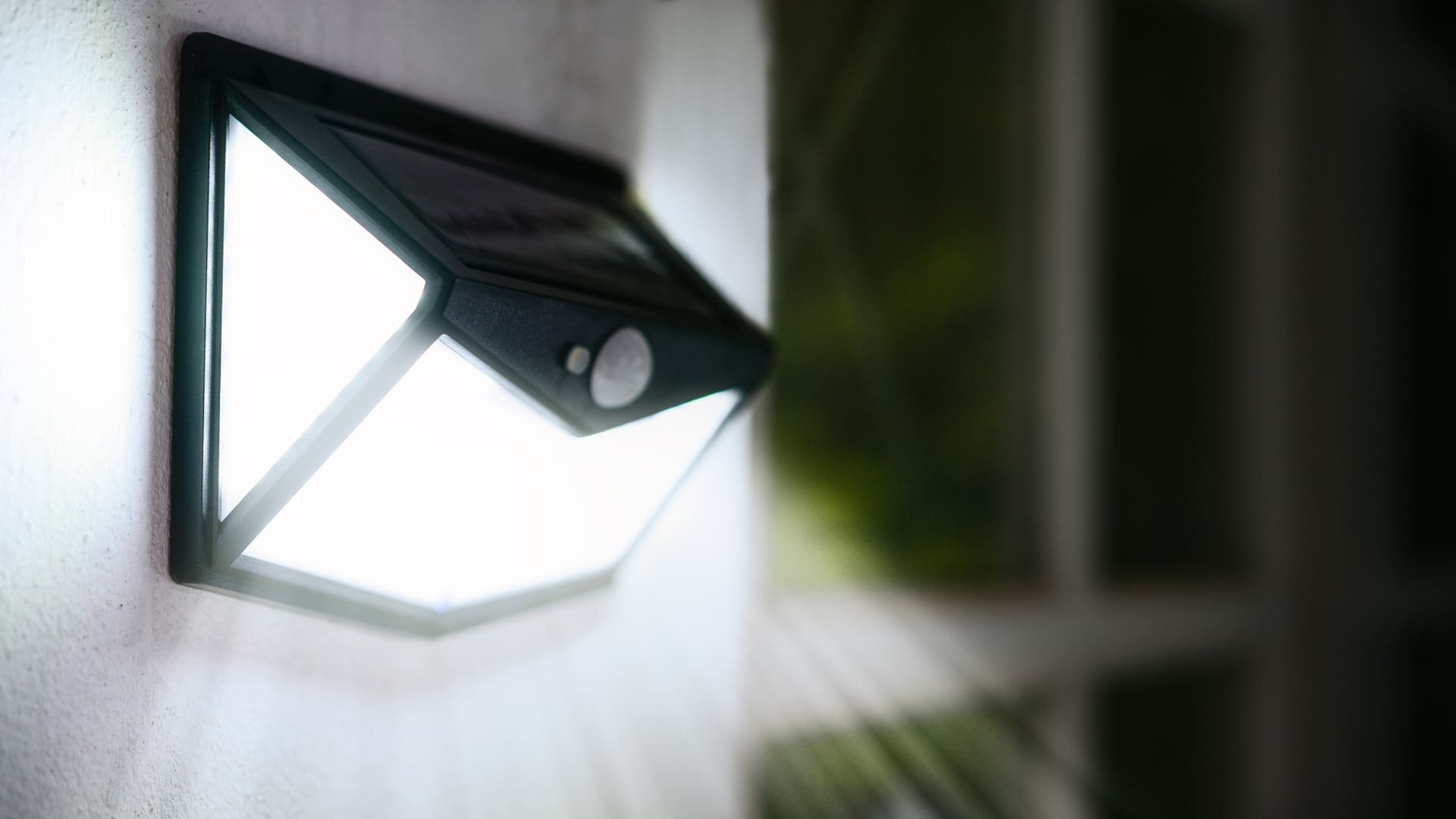 Wall-mounted outdoor light fixture emitting a bright light, with motion sensor near a blurred background.