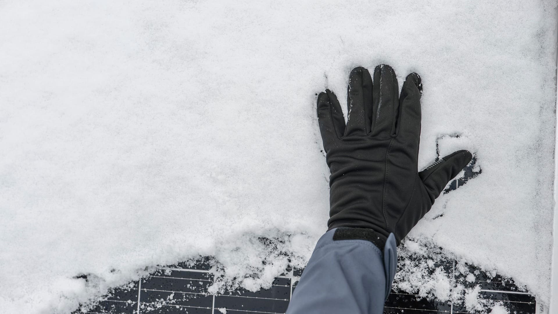 Gloved hand on snow-covered solar panel, partially cleared, highlighting the effects of snow on efficiency.
