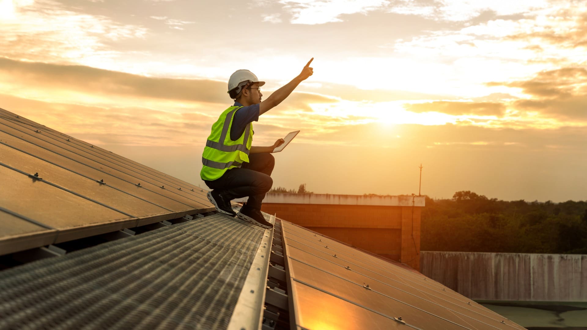 Worker in hard hat inspecting solar panels at sunset, holding tablet and pointing to the sky.