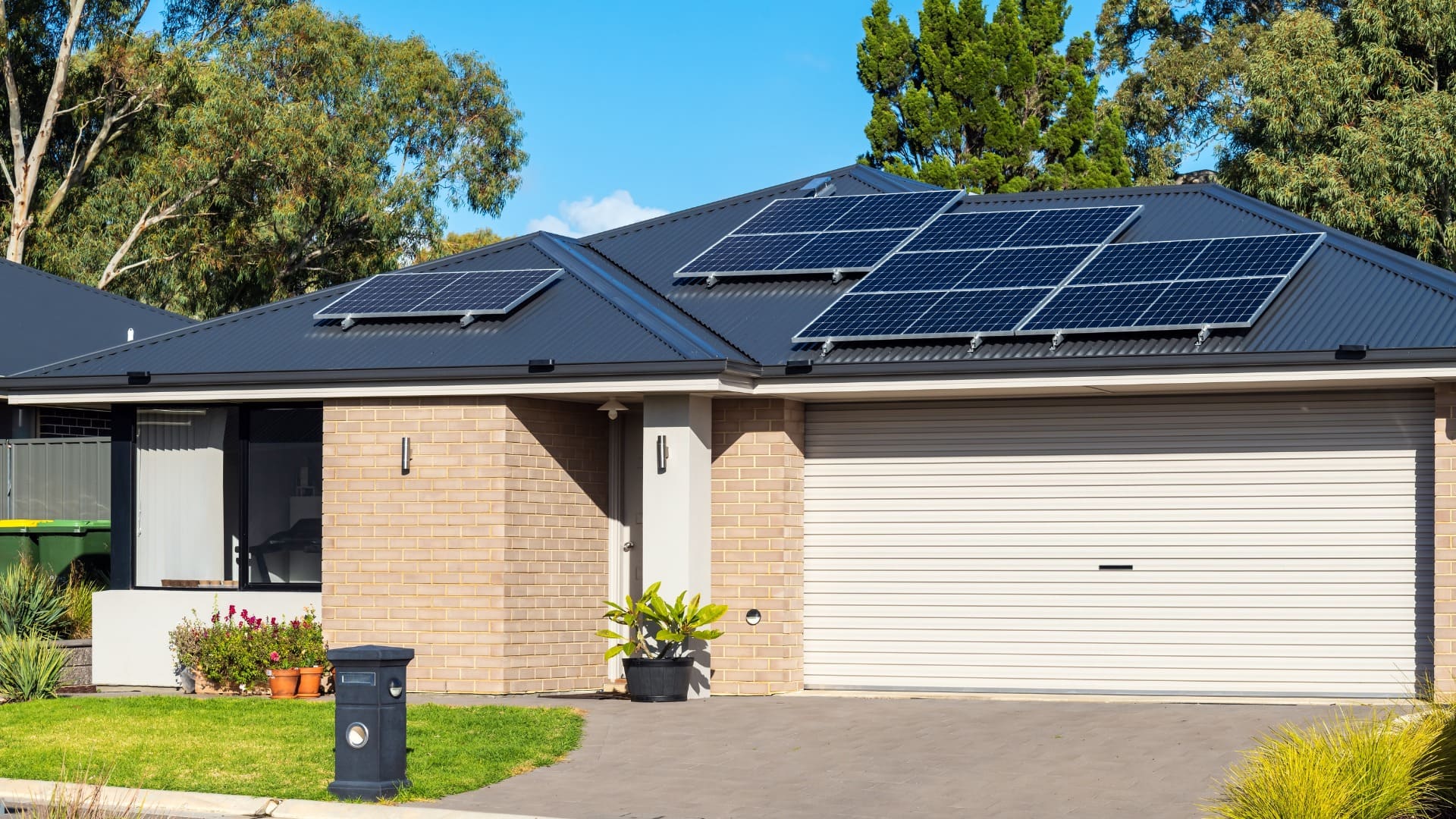 Modern single-story house with solar panels on roof, brick exterior, plants, and trees under blue sky.
