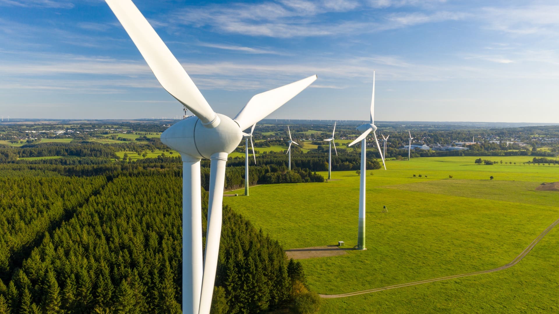 The image shows a wind farm with several wind turbines situated in a green landscape. The foreground features a close-up view of a wind turbine, while multiple other turbines are visible in the background. The wind turbines are positioned in an open field surrounded by patches of forest and a distant view of a town or village. The sky is clear with a few clouds, indicating a sunny day. This image is interesting and relevant as it depicts renewable energy sources, specifically wind power, which is an important aspect of sustainable energy solutions.