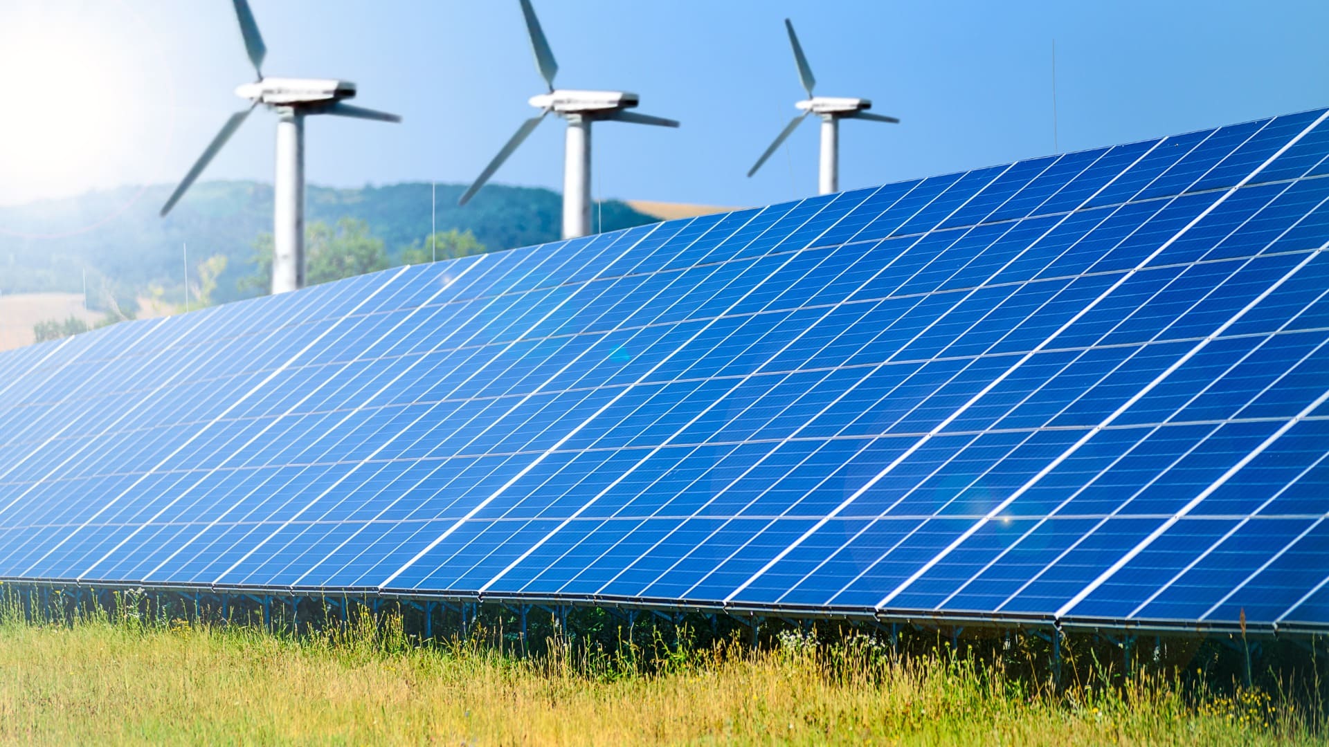 Field of solar panels with wind turbines on grassy hills, showcasing renewable energy sources in rural setting.