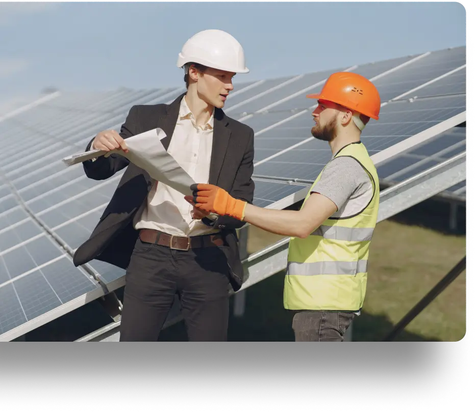 Two men stand in front of solar panels. One man in a suit and white hard hat holds a blueprint. The other man wears an orange hard hat, reflective vest, and gloves. They appear to be discussing a project. The sky is clear.
