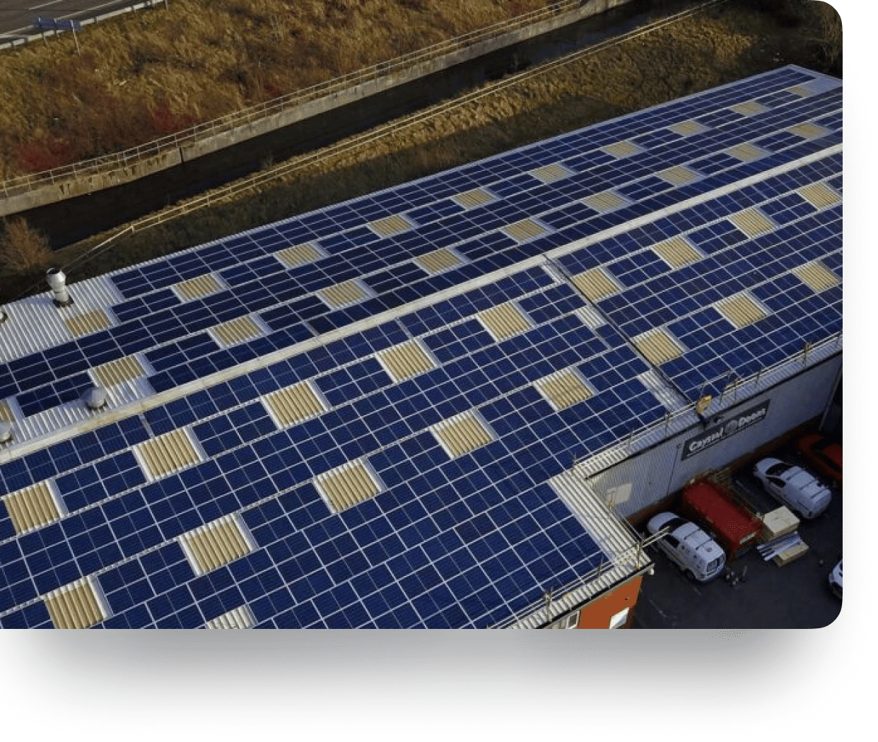 Rooftop of a large building covered with solar panels, with vehicles parked nearby and a grassy background.