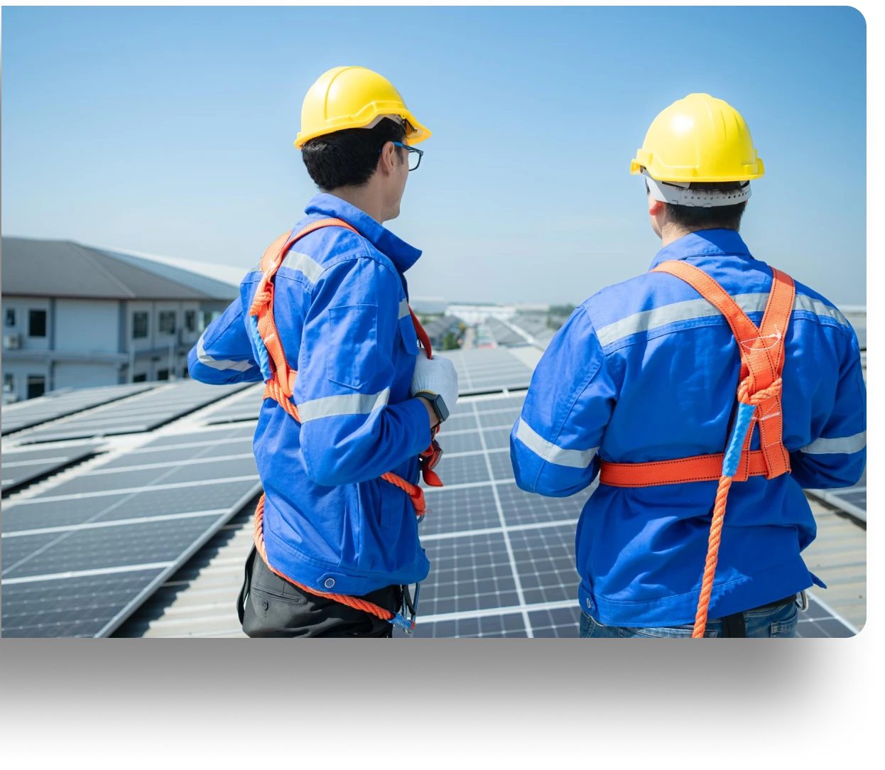 Two workers in uniforms and hard hats on a solar-panel-covered rooftop.