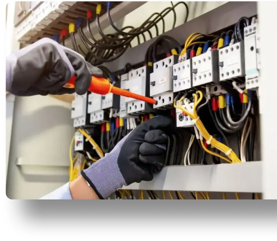 A person wearing gloves uses a screwdriver on an electrical panel filled with wires and circuit breakers.