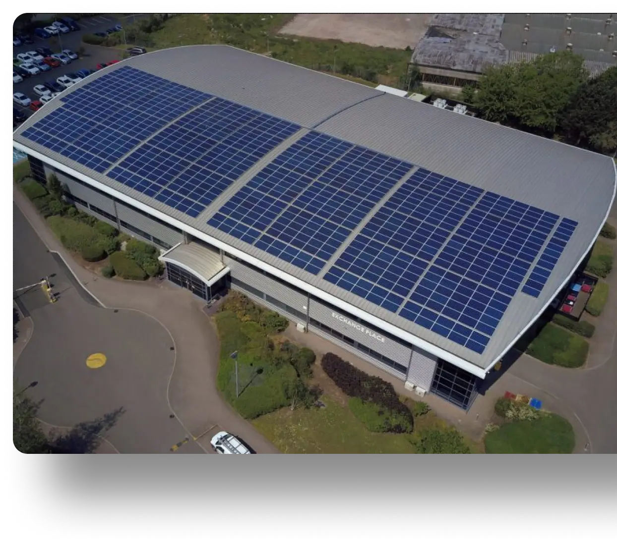 Aerial view of a large commercial building with a solar panel-covered roof, surrounded by greenery and parking areas.