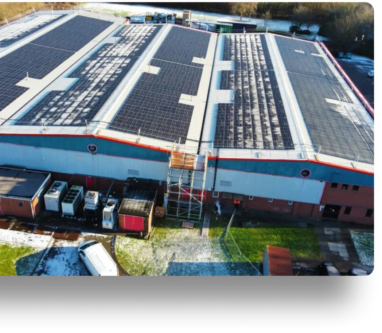 Large industrial building with solar panels on its roof, surrounded by equipment and patches of snow.