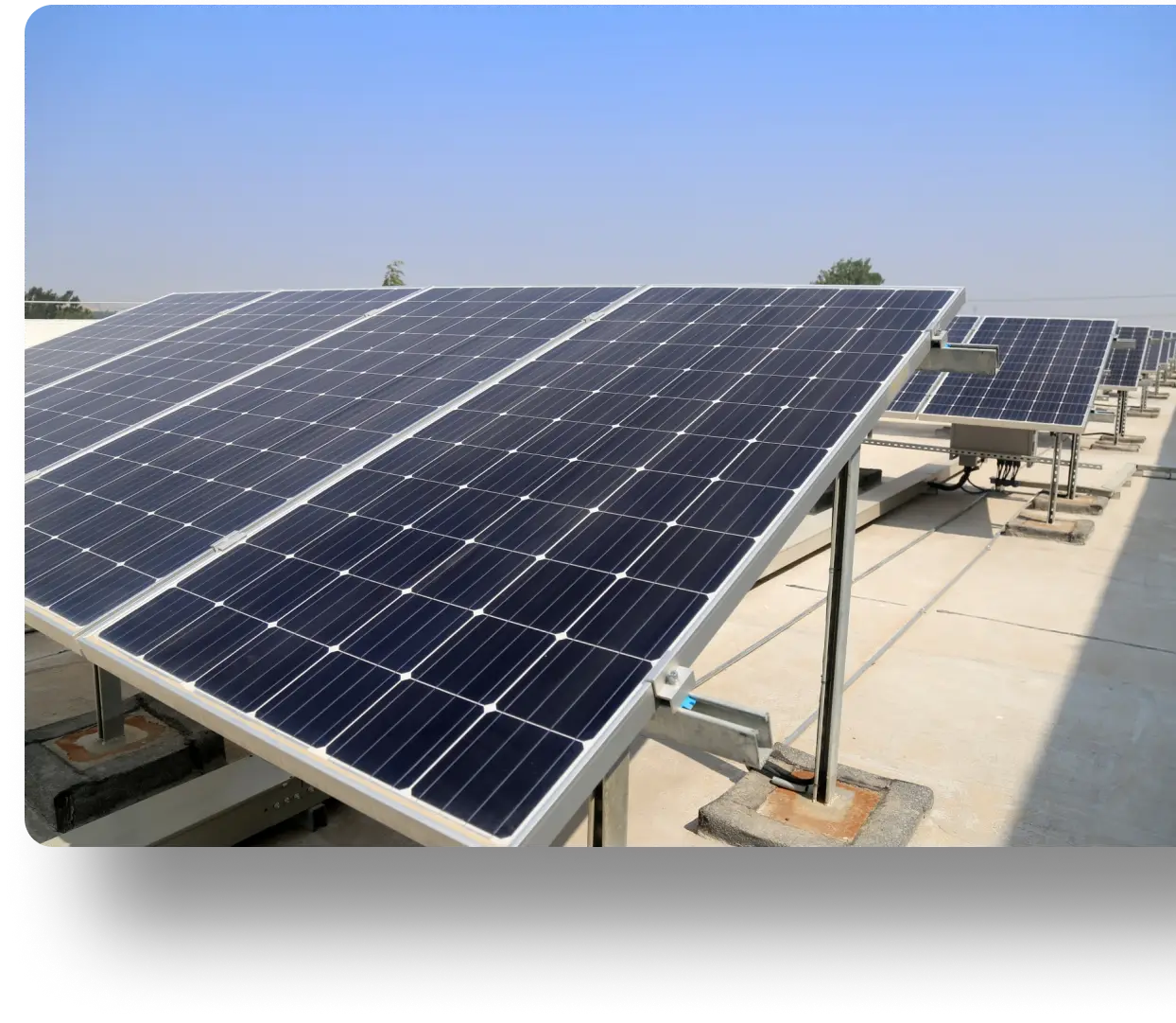Solar panels on a rooftop, angled towards the sky, with a clear blue backdrop. The panels are arranged in multiple rows, mounted on metal supports. The background shows a few trees on the horizon.