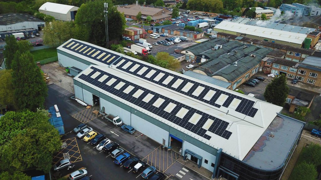 Aerial view of an industrial building with solar panels installed on its roof, surrounded by parked cars and trees.