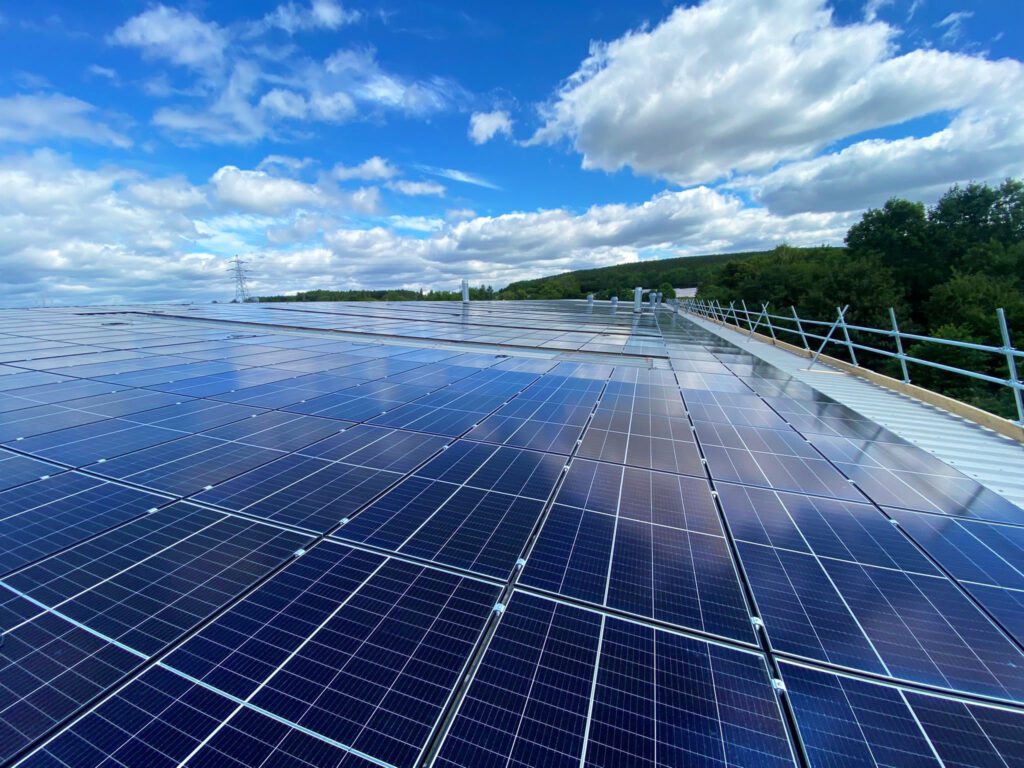 Rooftop solar panels arranged in neat rows, with a partly cloudy sky and trees visible in the background.