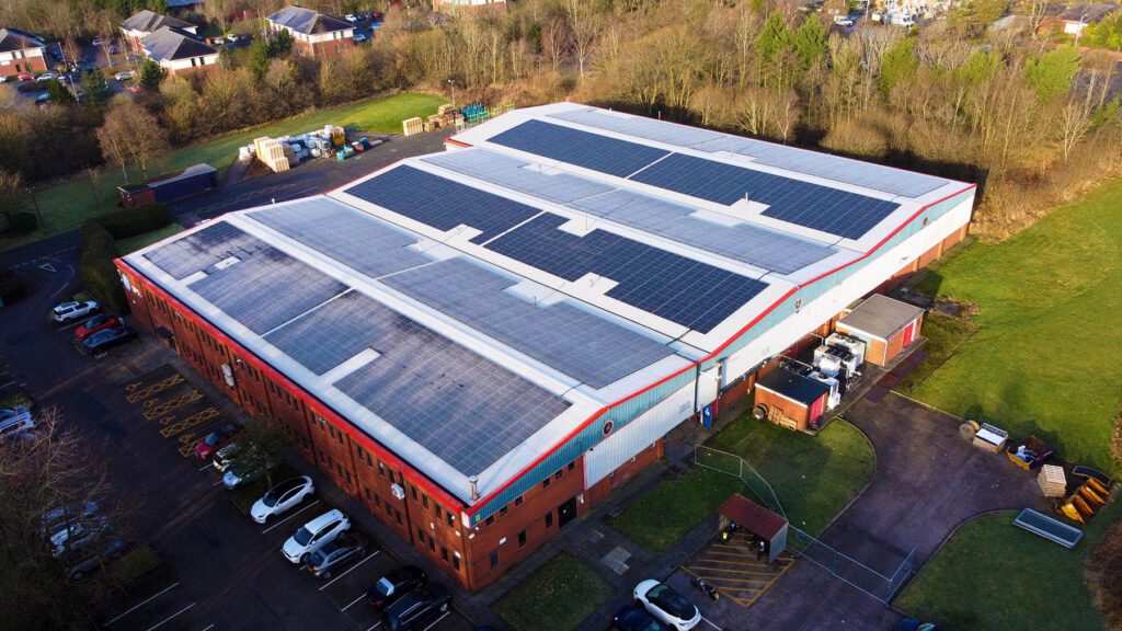 Aerial view of an industrial building with solar panels installed on its roof, surrounded by parking spaces and greenery.