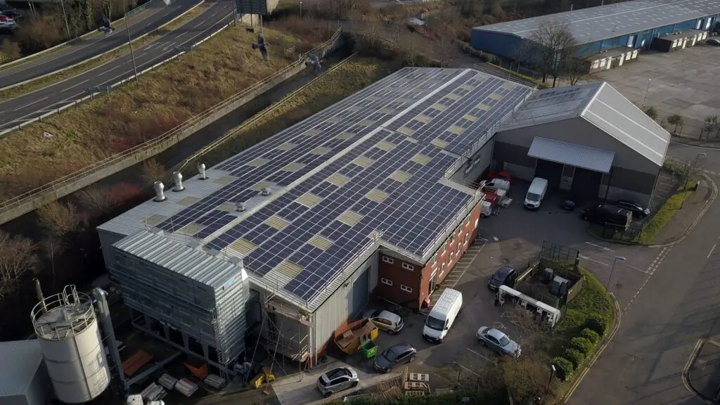 Aerial view of an industrial facility with solar panels on the roof, parked vehicles, and nearby greenery.