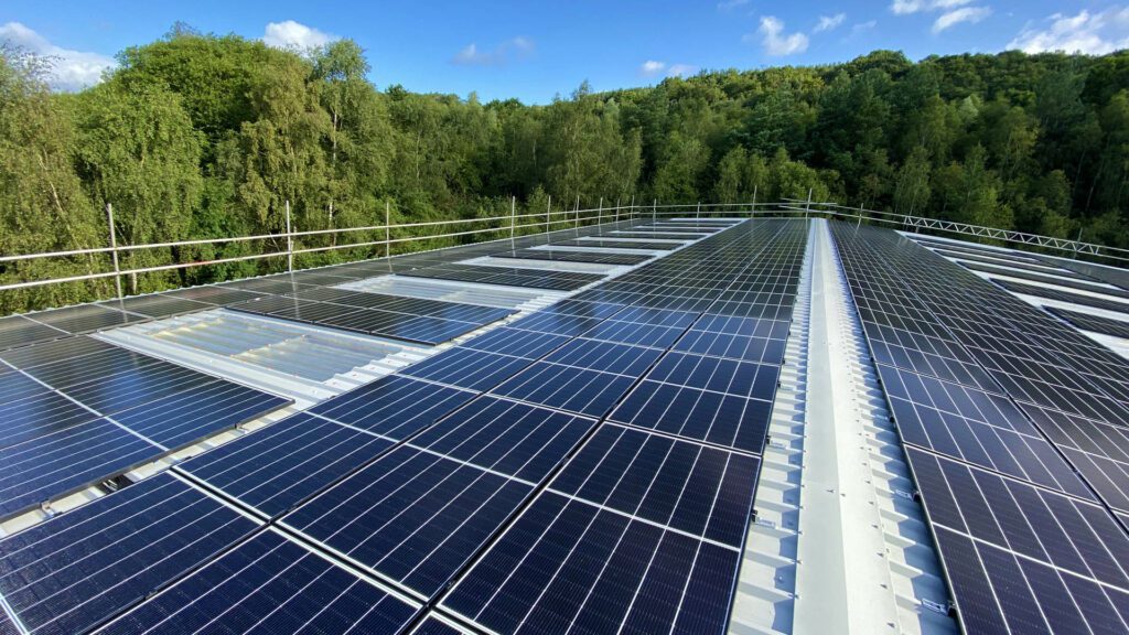 Aerial view of a rooftop solar panel system surrounded by lush green trees, under a clear sky with clouds.
