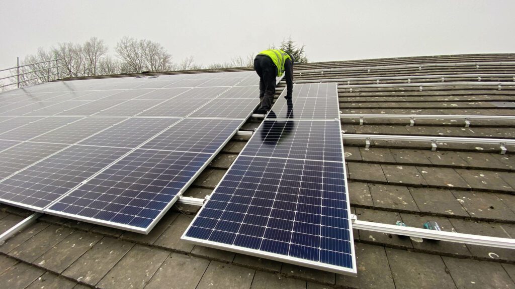 Worker in a high-visibility vest installing solar panels on a sloped roof, with mounting rails visible.
