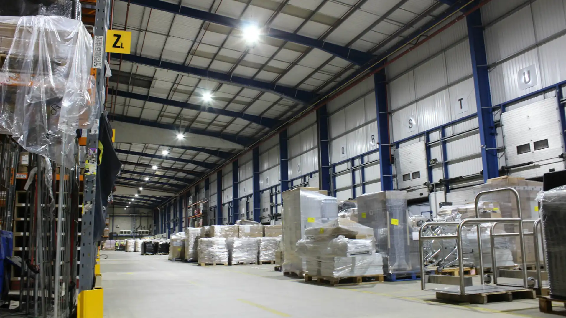 Warehouse interior with shelves, pallets, and high metal beam ceilings.