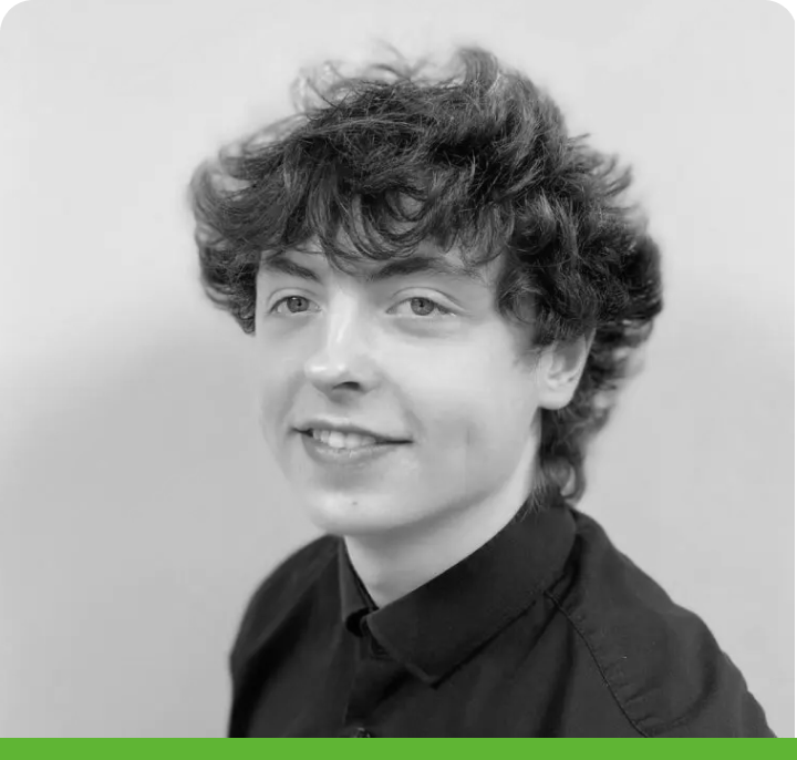 A person with curly hair smiles slightly, wearing a dark collared shirt. The black and white image has a green bar at the bottom, and the background is light-colored.
