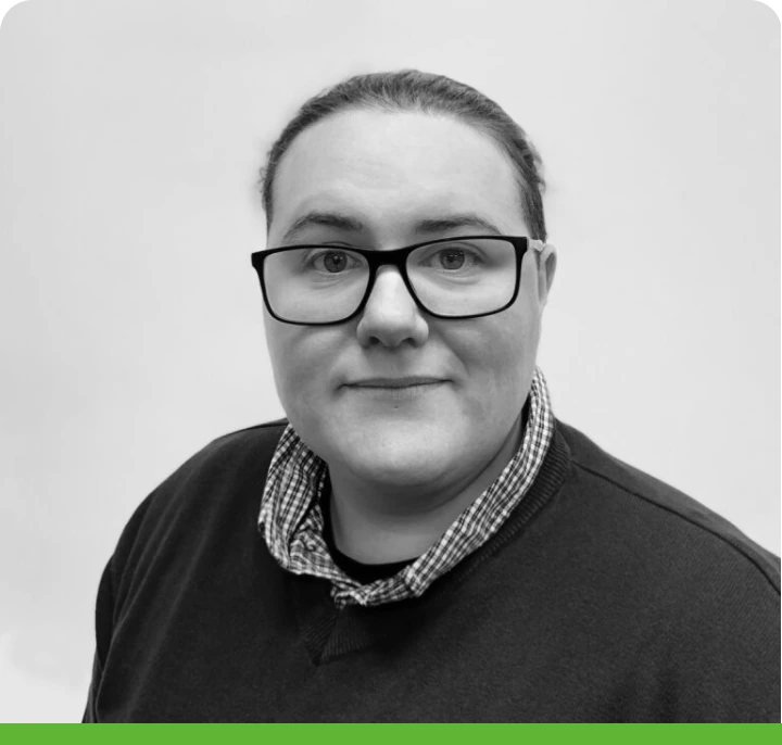 Person with glasses and short hair, wearing a dark sweater over a checkered shirt, looks at the camera. Background is plain with a green border at the bottom. Black and white image.