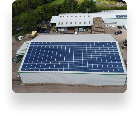 Aerial view of D&L Growers industrial building with a metal roof covered in solar panels. Nearby, another solar-paneled building is surrounded by trees. Vehicles and materials are arranged outside.