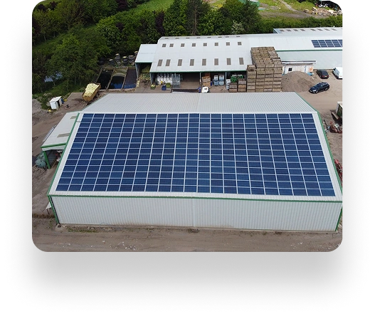 Aerial view of D&L Growers industrial building with a metal roof covered in solar panels. Nearby, another solar-paneled building is surrounded by trees. Vehicles and materials are arranged outside.