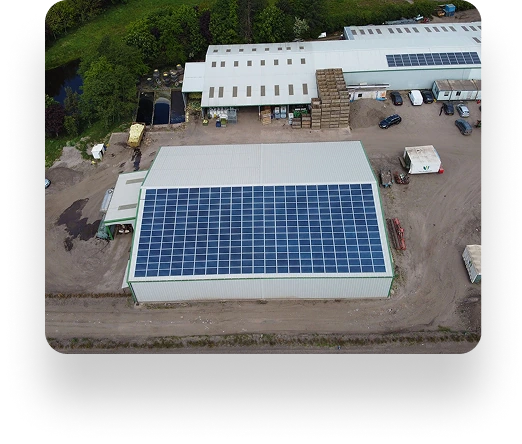 Aerial view of the D&L Growers industrial building with a solar-panel-covered roof. Surrounding it are dirt roads, parked trucks, smaller structures, greenery, and a nearby body of water.
