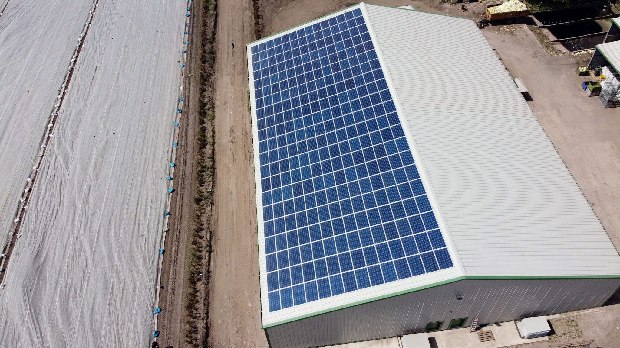 Aerial view of a building with solar-panelled roof beside fields, surrounded by roads and structures.