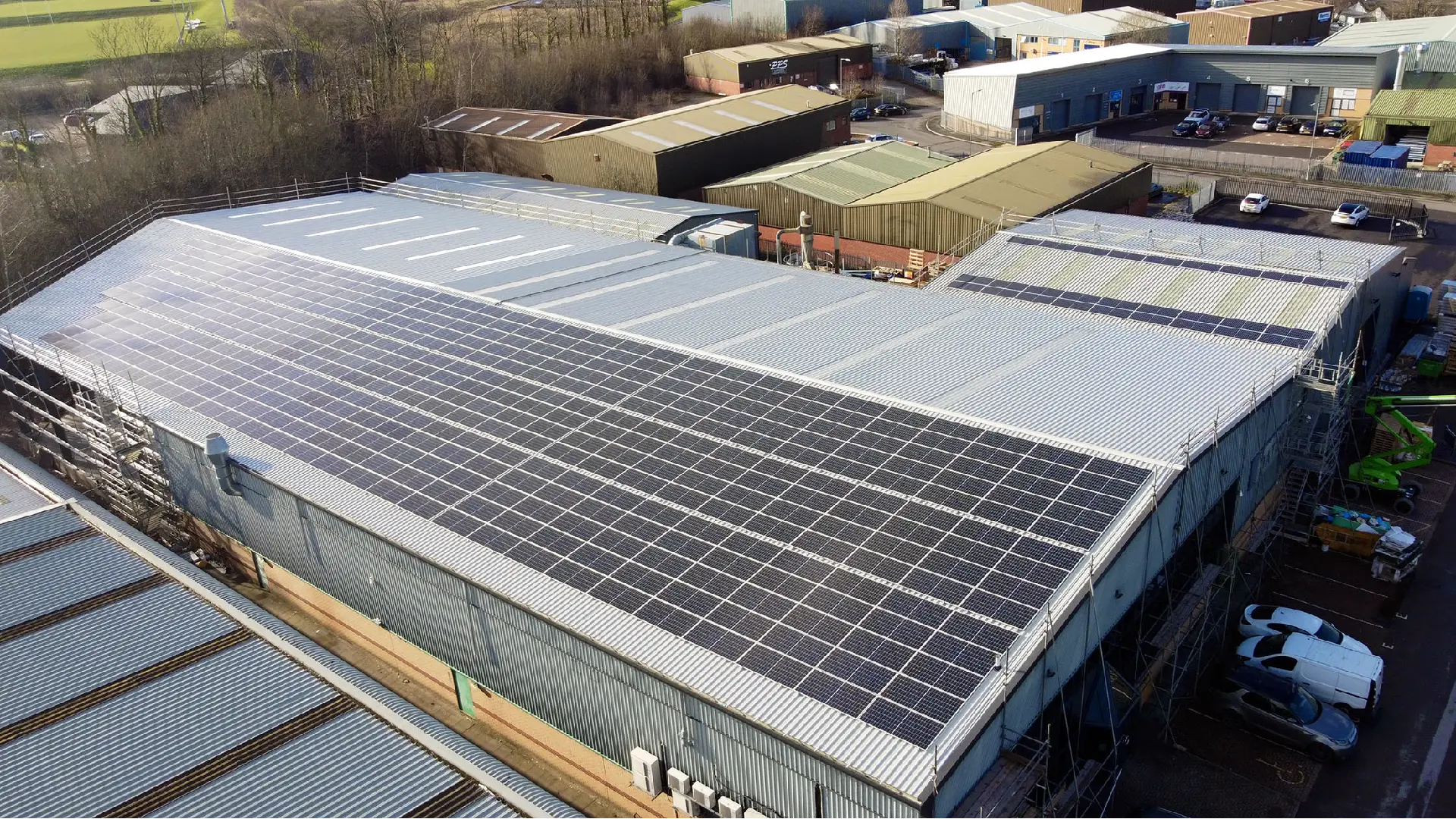 Aerial view of a large industrial building with a solar-panel-covered roof, surrounded by warehouses and offices. Trees line the property edge and few vehicles are parked nearby.