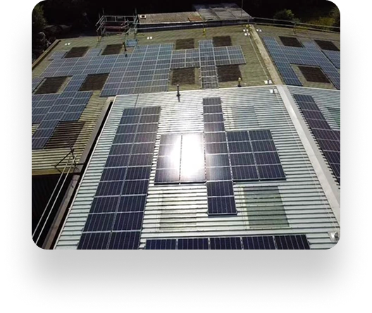 A rooftop covered with solar panels of various sizes, installed by Dean Group. Sunlight reflects off the central panel, while scaffolding is visible in the background. The sky is not in view.