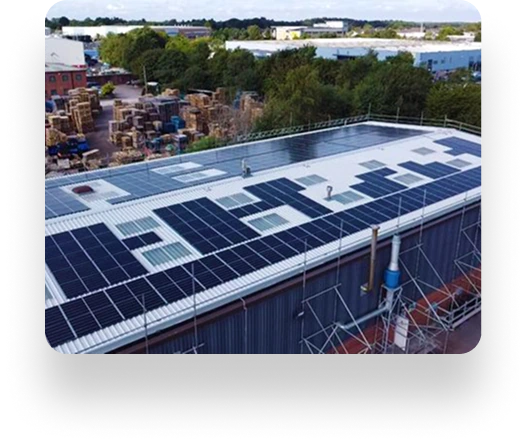 Aerial view of a flat industrial rooftop with solar panels, installed by Dean Group. Workers inspect panels. Trees and industrial storage areas surround the building.