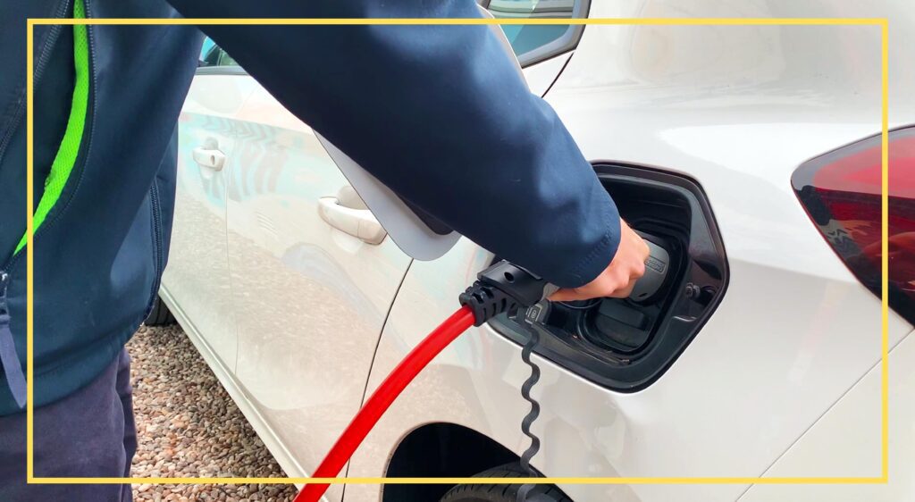 Person plugging a red charging cable into a white electric vehicle, wearing a dark jacket with a green stripe.