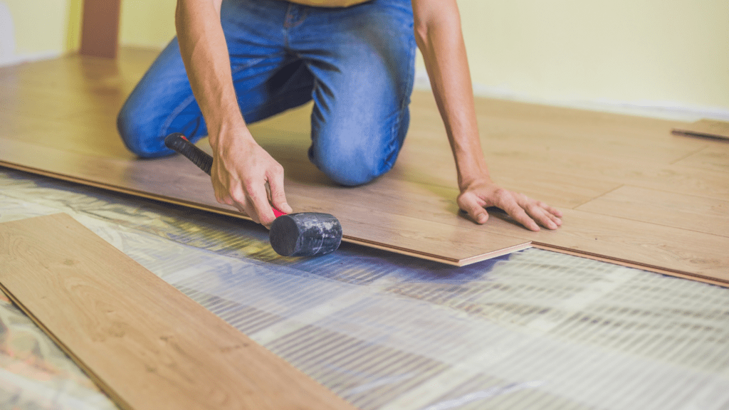 Person installing wooden flooring with a hammer, kneeling on the floor, securing planks in place.