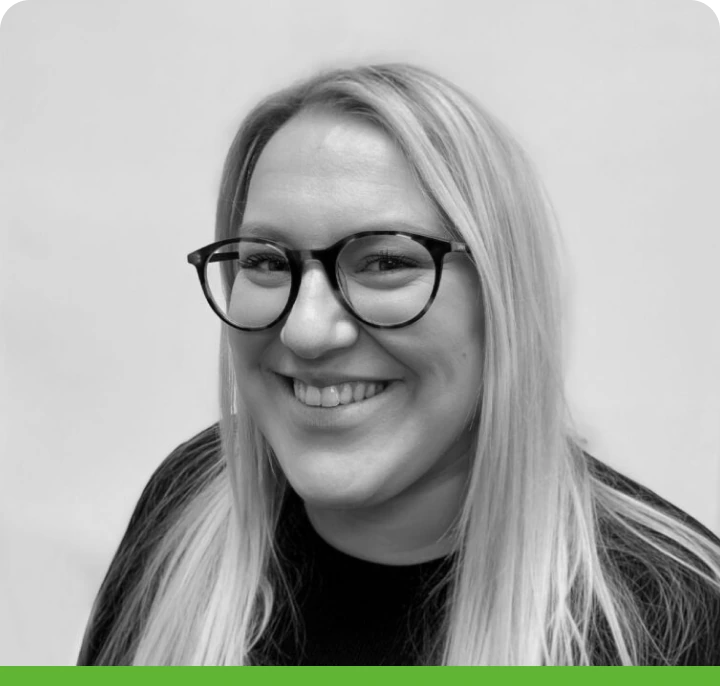 A person with long, light hair and glasses is smiling at the camera, wearing a dark top. The black-and-white image features a rounded green border at the bottom.