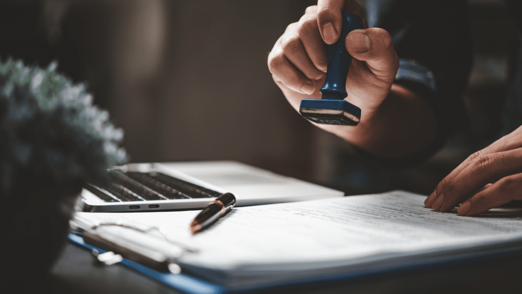Person pressing a blue stamp onto a document on a clipboard, with a pen and a laptop visible nearby.
