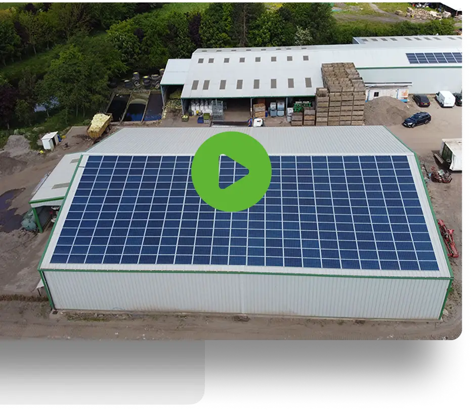 Aerial view of a large industrial building with solar panels covering its roof. A play button icon is superimposed on the image, suggesting a video. Surrounding the building are trees, other structures, and stacks of materials.