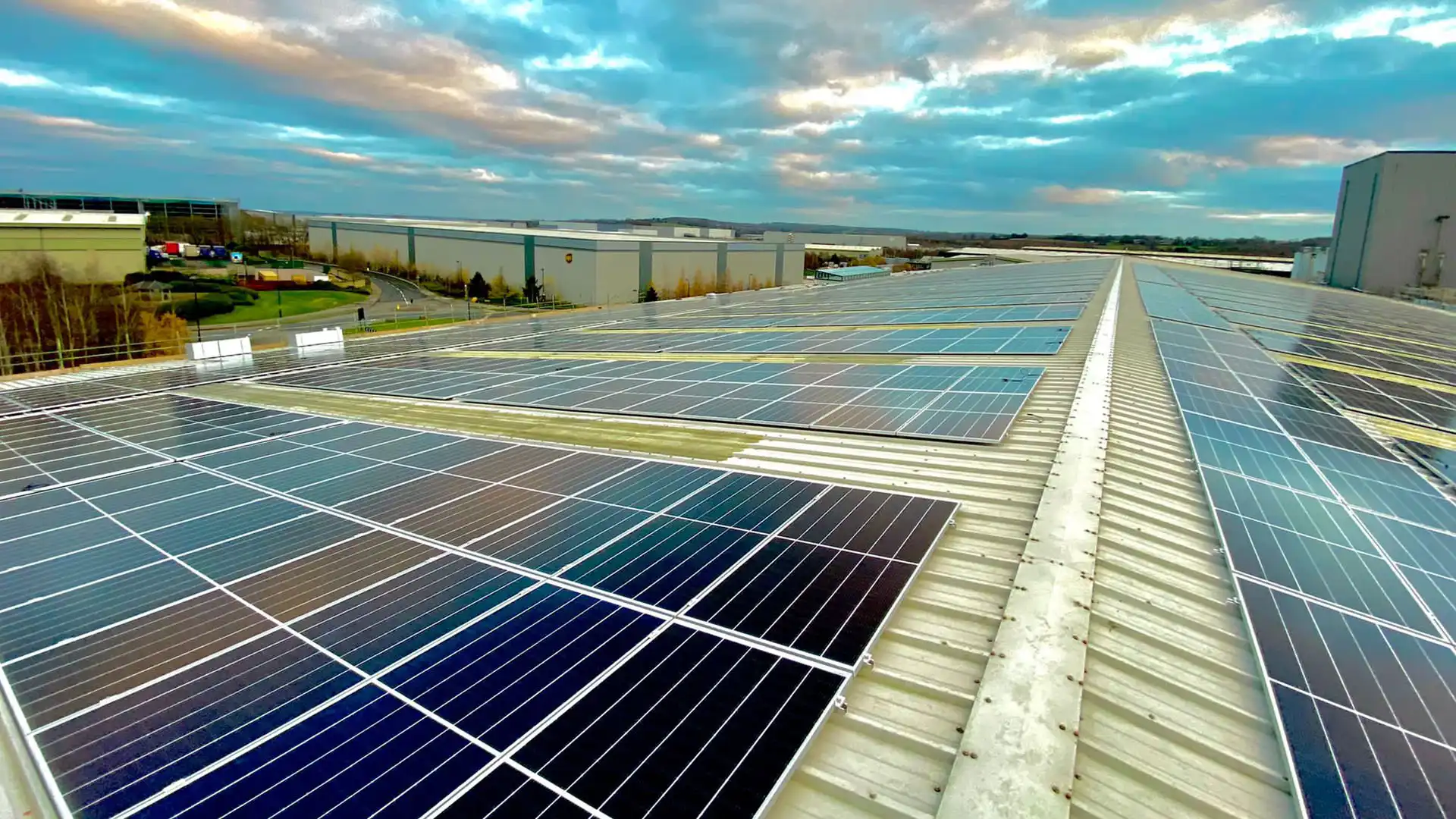 Rooftop with Instarmac solar panels stretching into the distance under a partly cloudy sky. Reflections create vibrant colors; industrial buildings and greenery line the horizon.