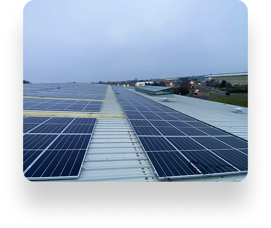 Rows of solar panels cover a flat rooftop under an overcast sky. An Instarmac roadway, fields, and buildings are visible in the distance. The aligned panels reflect minimal sunlight.