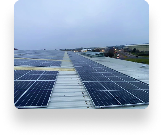 Rows of solar panels cover a flat rooftop under an overcast sky. An Instarmac roadway, fields, and buildings are visible in the distance. The aligned panels reflect minimal sunlight.