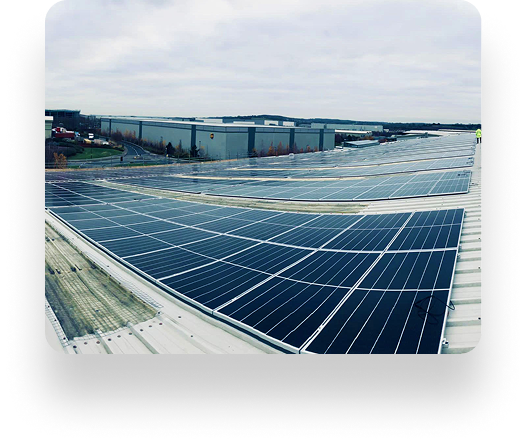 Rooftop solar panels cover a large industrial building beneath a cloudy sky. The Instarmac installation is orderly, capturing renewable energy. Nearby buildings and a distant treeline complete the view.