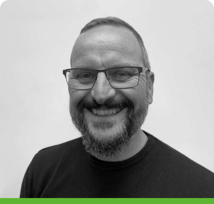 A man with glasses and a beard smiles at the camera. He is wearing a dark shirt against a plain background. The photo is black and white with a green border.
