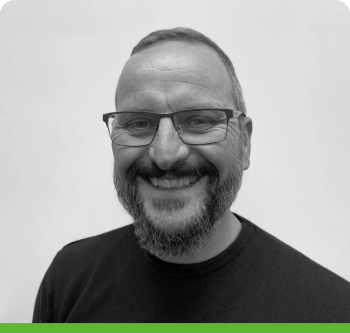 A man with glasses and a beard smiles at the camera. He is wearing a dark shirt against a plain background. The photo is black and white with a green border.