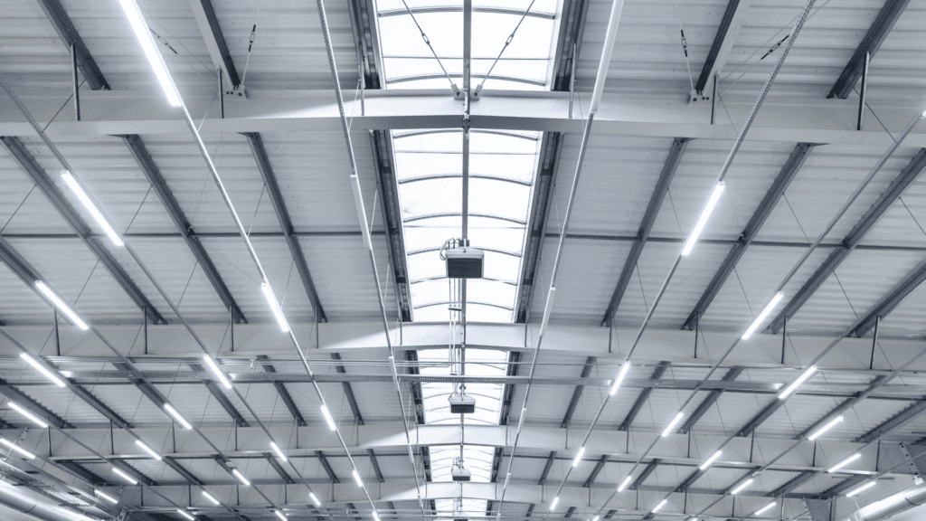 Interior ceiling of an industrial building with metal beams, long fluorescent lights, and a central skylight.