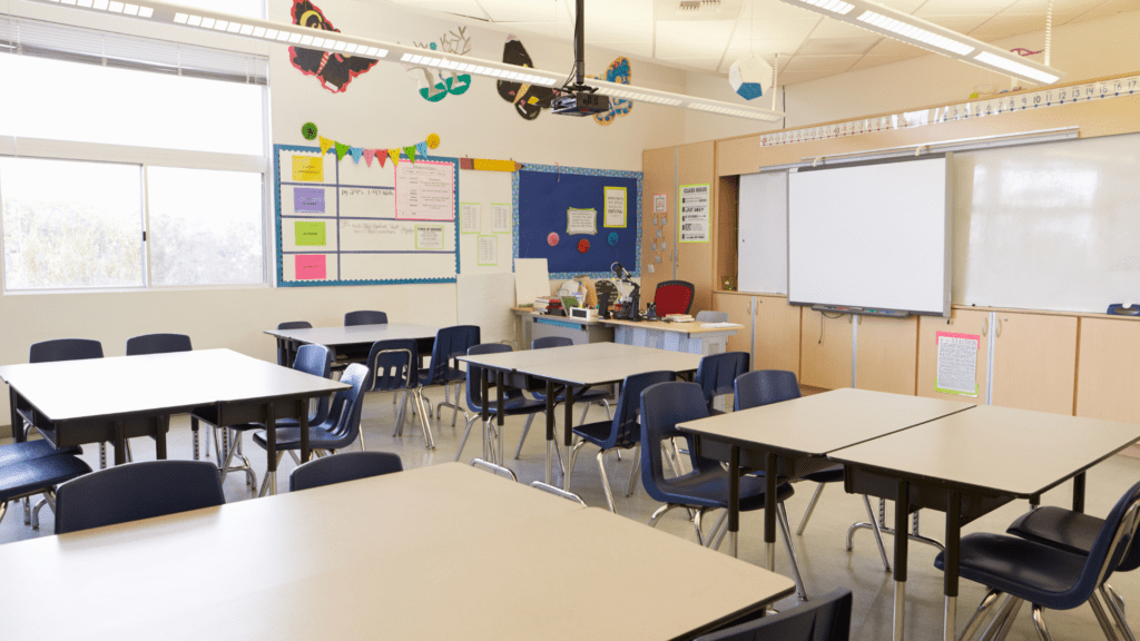 Classroom with tables and chairs arranged in groups, educational posters on walls, and a whiteboard upfront.