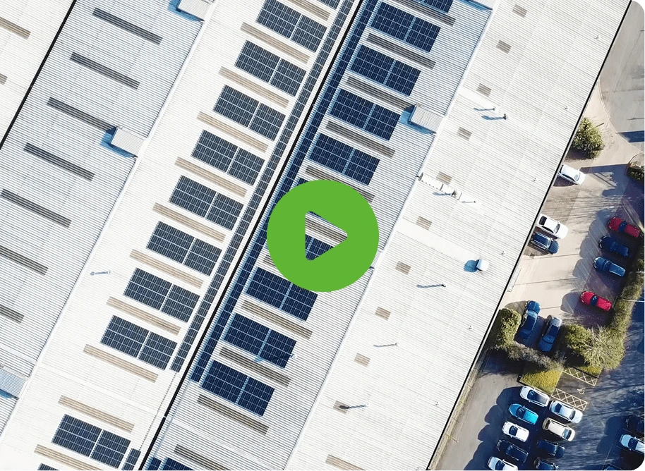 Aerial view of a large white rooftop with solar panels, highlighting Laltex Groups innovation. A green play button overlay is centered. Bottom right shows a busy parking lot lined with trees.